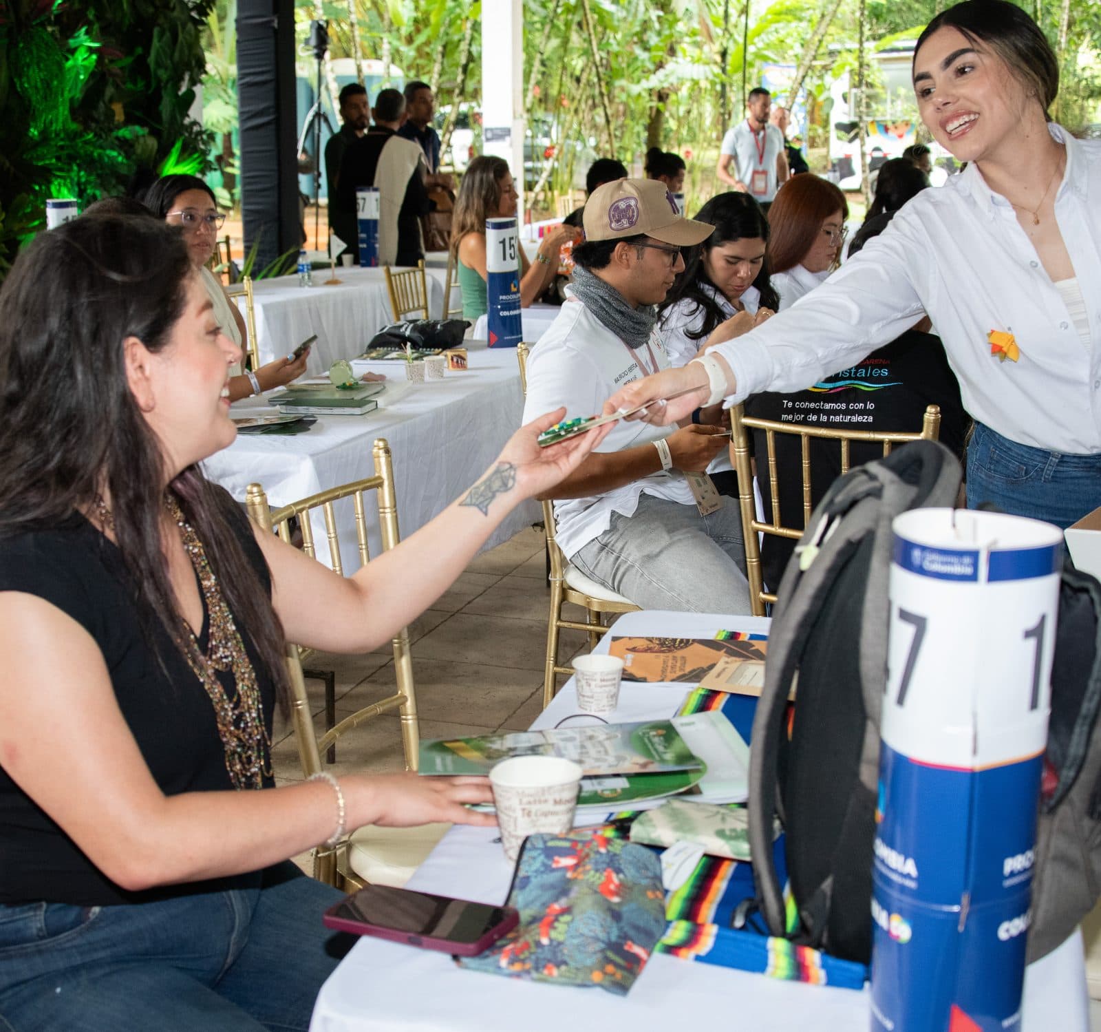 Una mujer de camisa blanca conversa con una visitante sentada a una mesa, en un salón lleno de mesas con material de turismo.
